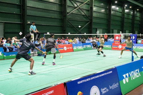 Badminton match in progress with players on a court, surrounded by spectators and Yonex advertisements.